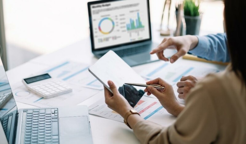 Woman gesturing at tablet with a stylus to show her coworker something. Papers scattered on desk in background plus a computer screen showing charts.