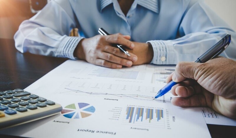 Coworkers seated at a table with several papers that have charts on them. One colleague points with a pen at a page while the other coworker observes.