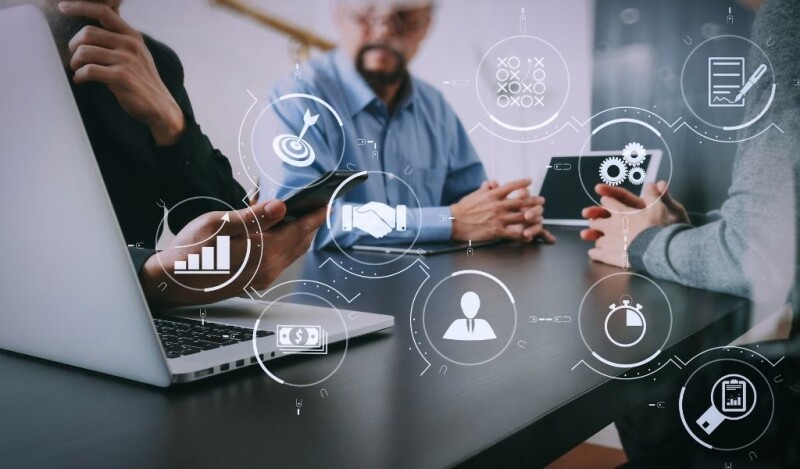Colleagues in a meeting with icon imagery overlaid containing: target, graph, clock, cash, gears