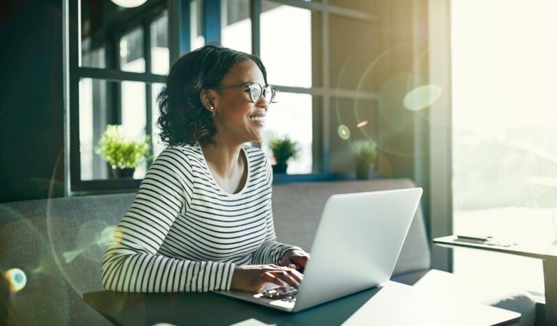 Woman smiling at laptop while working while the sun shines in