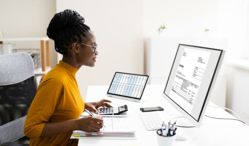 Woman smiling at computer screen while using a notepad and calculator. Computer shows an invoice.