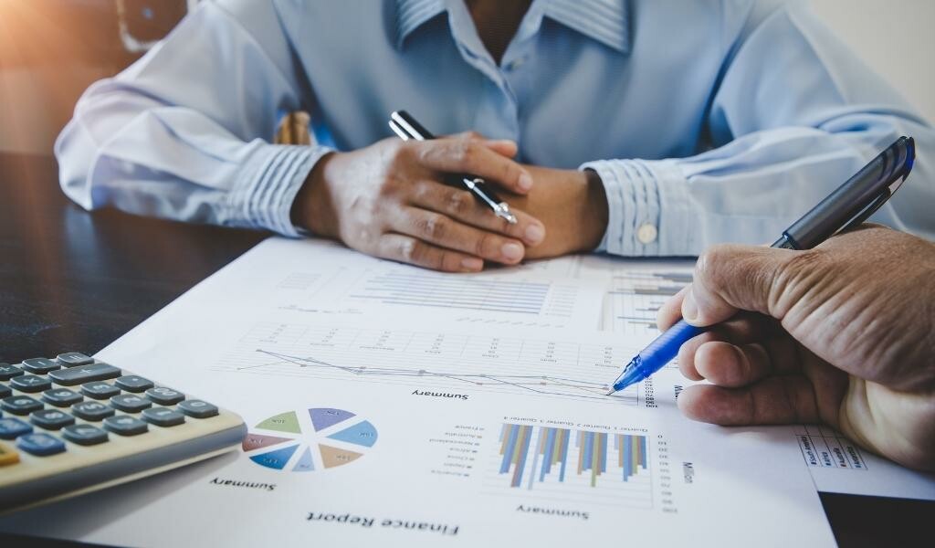 Coworkers seated at a table with several papers that have charts on them. One colleague points with a pen at a page while the other coworker observes.