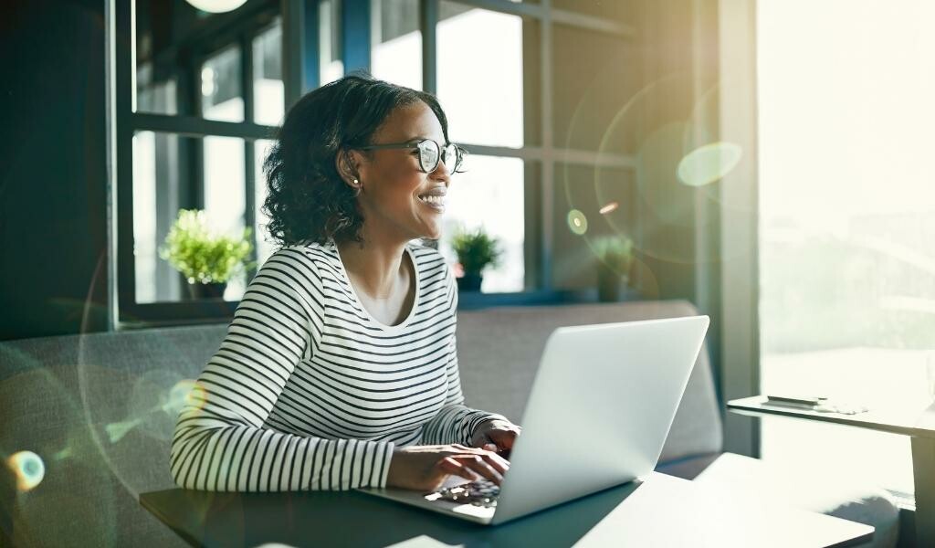 Woman smiling at laptop while working while the sun shines in