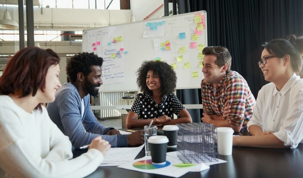 Colleagues working together at a table with a whiteboard in the background depicting ideas
